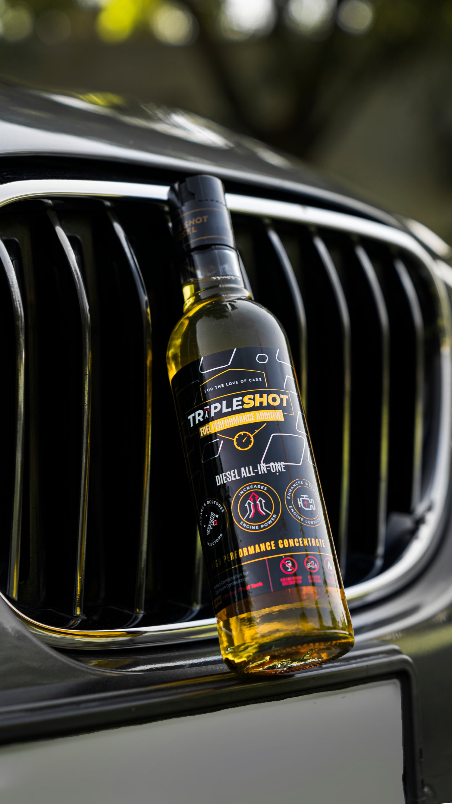 Tripleshot Diesel All-in-One Fuel Performance Additive bottle placed leaning against the front grille of a grey car, with a focus on the bottle label and yellow liquid inside.