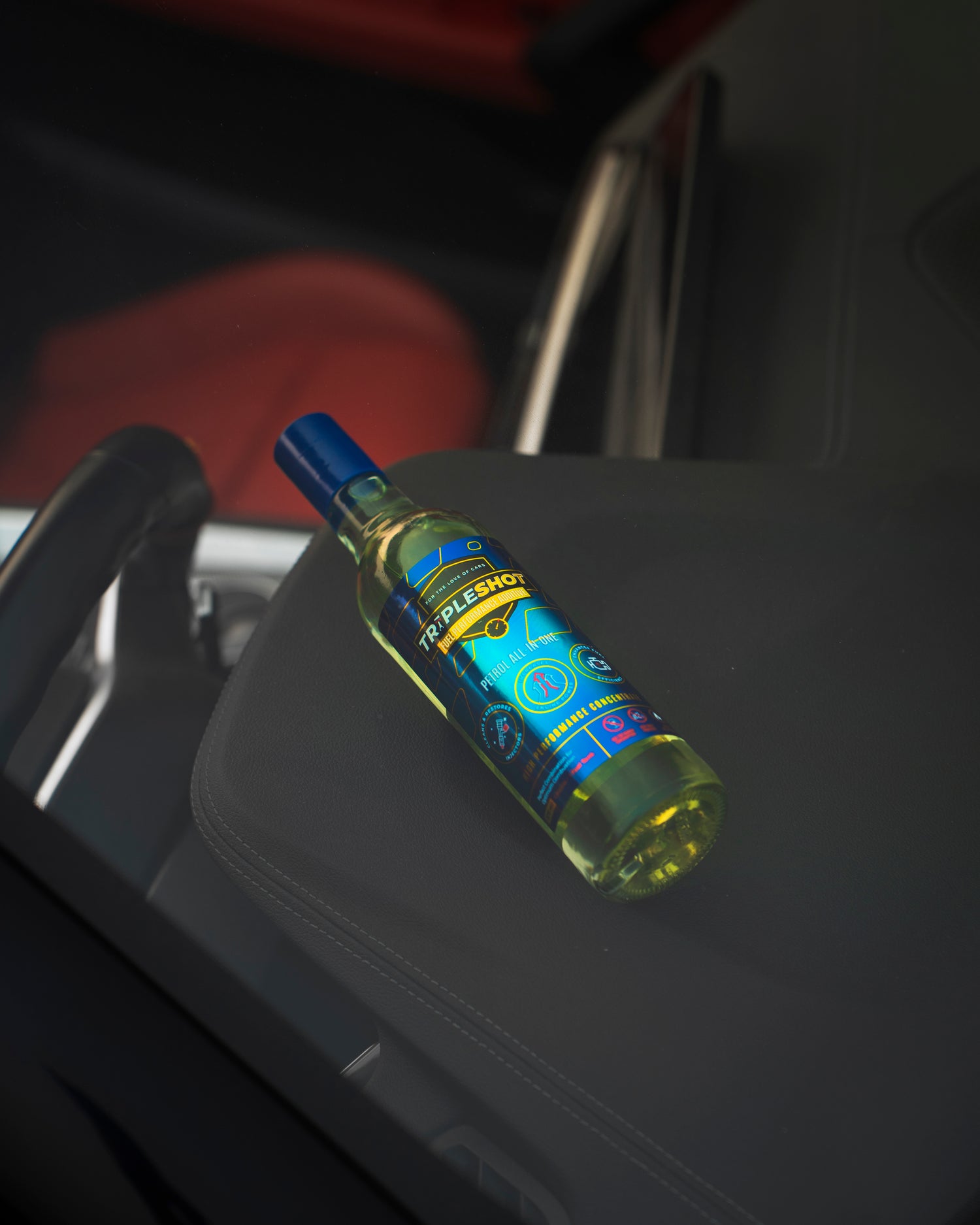 Tripleshot Petrol All-in-One Fuel Performance Additive bottle with blue cap and label, placed on the armrest inside a car, with part of the steering wheel and red car seat visible in the background.